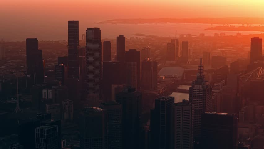 Wide aerial panorama of Melbourne skyline during fiery sunset over city, drone ascend overview looking down