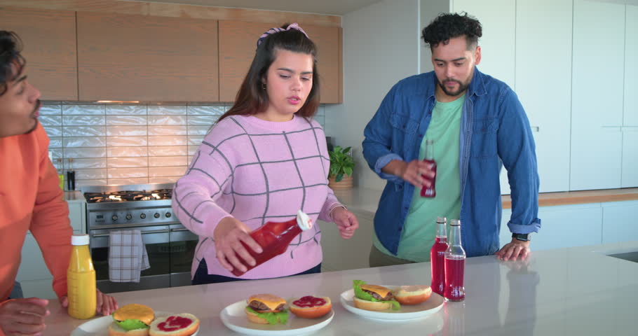 Diverse friends assembling burgers for meal prep after ketchup squeeze, shaking mustard at island. Collaboration, casual, vibrant, social, cuisine, friendship, modern