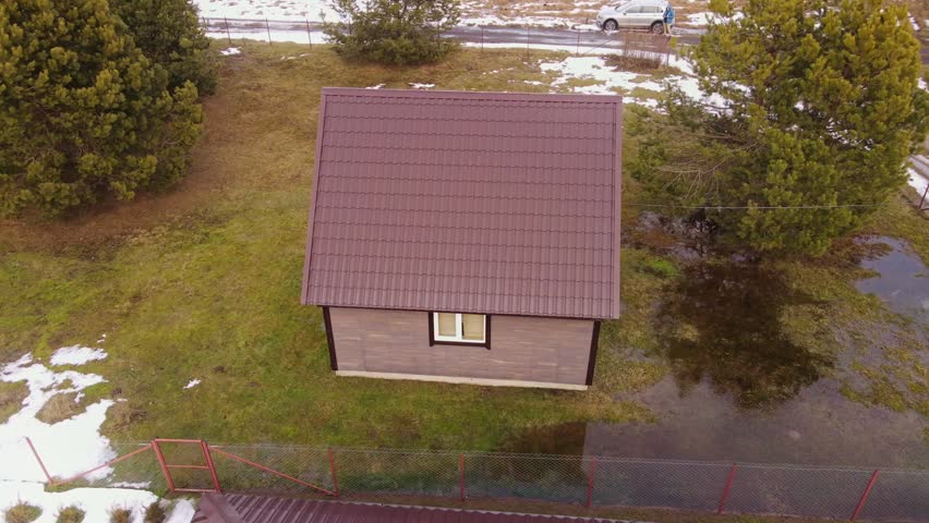 Aerial view of a Belarus homestead, brown roof cottage, greenhouse, fenced plots, muddy lane, striped fields of snow and growth, conifers, car and person, overcast light