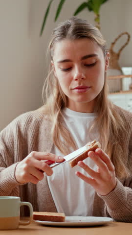 A young woman in her cozy kitchen spreads peanut butter on toast, taking a delightful bite. She savors a healthy breakfast with a warm cup of tea, embracing a serene morning routine at home.