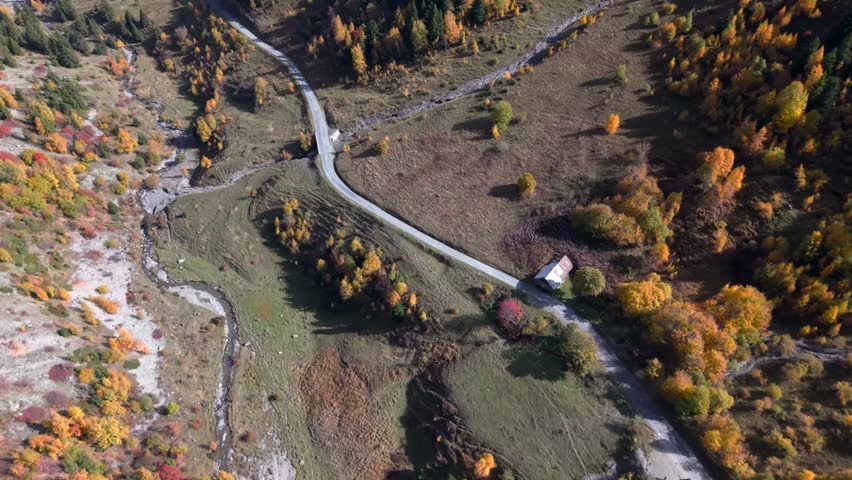 Aerial view of winding mountain road and river near Col du Glandon in the French Alps. Scenic autumn landscape with alpine valley, colorful trees and peaceful countryside.