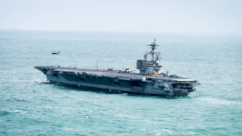 Large Aircraft Carrier Sailing on the Ocean Under Hazy Sky with Helicopter Flying Above and Personnel on Deck.