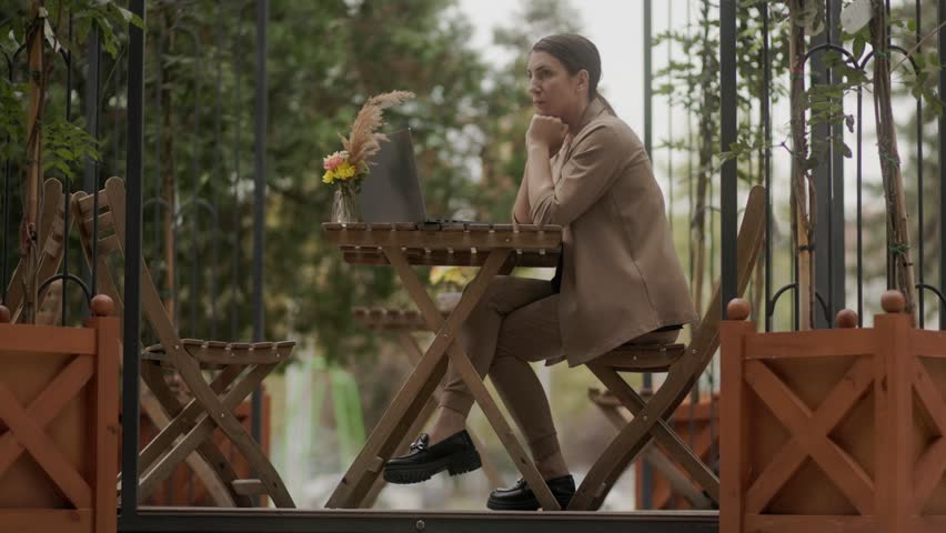 The image captures a thoughtful brunette woman in business attire working remotely, sitting on a terrace with her laptop, showing serious focus and determination.