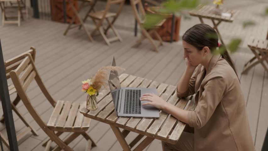 A young, serious businesswoman sits at an outdoor table, typing on her laptop. Her fatigued expression and posture suggest a long day of remote work.