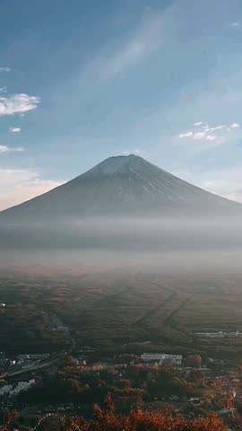 Mount fuji stands prominently at the center of the composition, its symmetrical volcanic cone rising above a wide valley layered in soft mist. Light snow traces the upper slopes, emphasizing the mountain’s ridges and natural contours. A pale blue sky with scattered white clouds stretches overhead, creating a calm atmospheric backdrop. Below the mountain, a dense expanse of forest and small structures spreads across the landscape, partially veiled by low morning haze. The scene highlights the iconic silhouette of mount fuji, the gentle gradation of light across the slopes, and the layered depth formed by mist, valley, and sky.