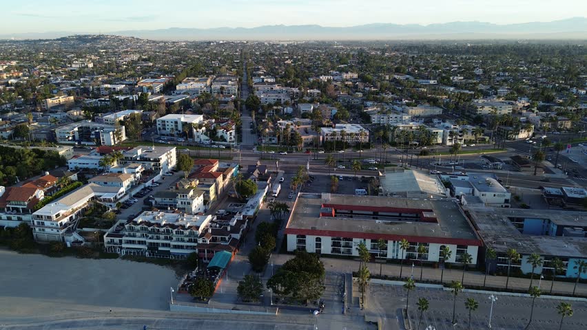 Backward flyover view Beachfront residential edge by Belmont Shore north of pier, East Ocean Blvd connects dense residential blocks to coast, lifeguard tower positioned along sand, Long Beach, CA. USA
