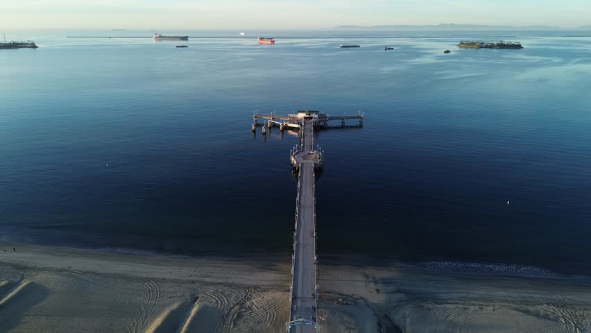 Pier and open-water span over Belmont Veterans Memorial extend toward harbor channel with anchored vessels visible beyond. Calm water, soft reflections, warm sky gradients, Long Beach, CA. USA