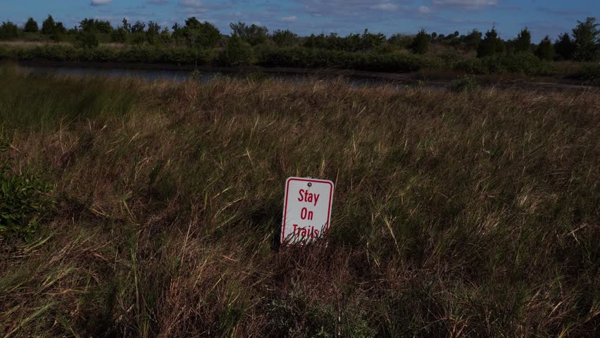 A field with very short vegetation has a sign to stay on the trails. In the background, one can see water, trees, and a sky with clouds.