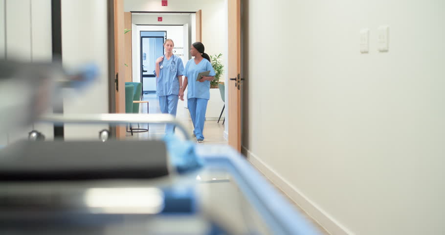 Diverse female nurses entering doorway in scrubs, walking and consulting tablet for care. Healthcare, teamwork, hospital, corridor, device, gurney, review