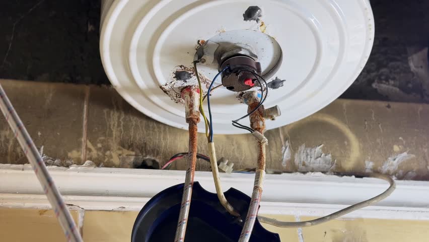 Close up of old electric water heater underside showing rusted pipes, exposed electrical wiring, and corrosion buildup. Indoor plumbing and heating system in poor maintenance condition.