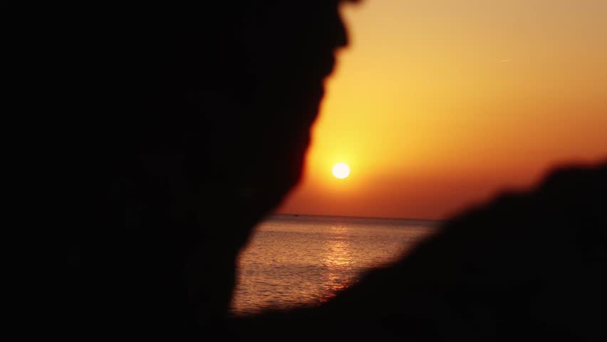 Sunset over Ibiza sea framed by blurred rocks with focus on golden sun and calm water horizon.