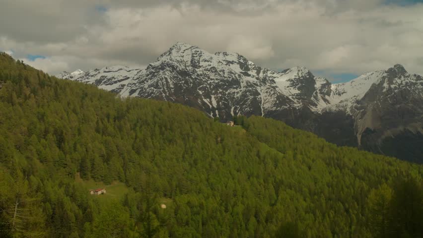 Rugged snow dusted alpine mountain peaks and dense green conifer forest landscape in Switzerland. Vast wilderness vista captures beauty of Graubunden region during summer season.