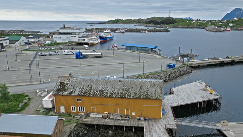 Drone view of harbor area in Lofoten Islands, Norway. Quiet norwegian fishing port with boats, warehouses and Arctic coastal scenery under cloudy sky.