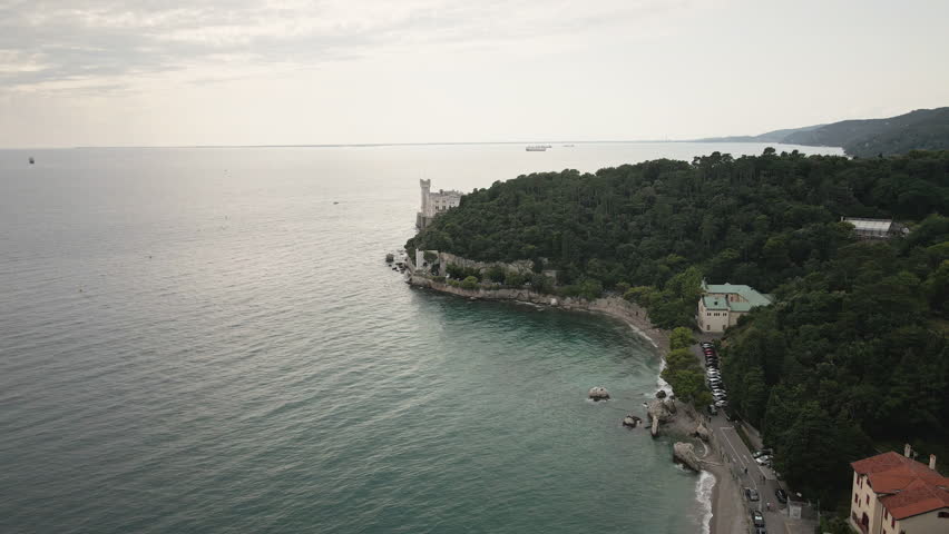 Cinematic 4K UHD drone flies forward along Trieste’s Adriatic coast toward Miramare Castle, revealing a forested headland and calm sea for travel ads, openers.