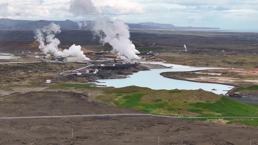 A geothermal powerplant in Iceland creates energy to use for the country and pumps out large plumes of steam into the sky near a mineral blue lake