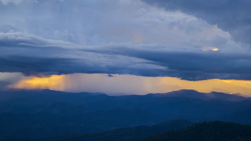 Dramatic timelapse zoom in of storm clouds over mountain range with golden sunset light breaking through horizon.