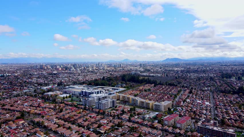 Aerial view of residential neighborhoods with terracotta roofs and modern apartment complexes spreading toward city skyline and Andes mountains under blue sky with white clouds. Santiago, Chile