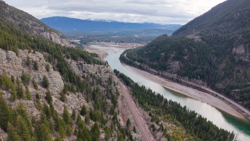 Aerial drone view of a turquoise river winding through a valley with rocky cliffs and evergreen forests on one side and open farmland and mountains on the other near Whitefish, Montana