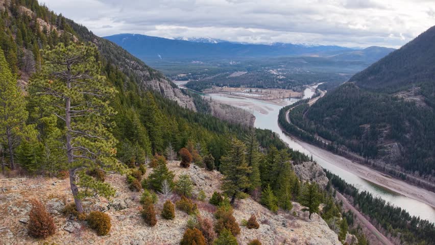 Aerial view of a river valley from a rocky hillside covered in evergreen trees, capturing the winding Flathead River and distant mountains near Whitefish, Montana