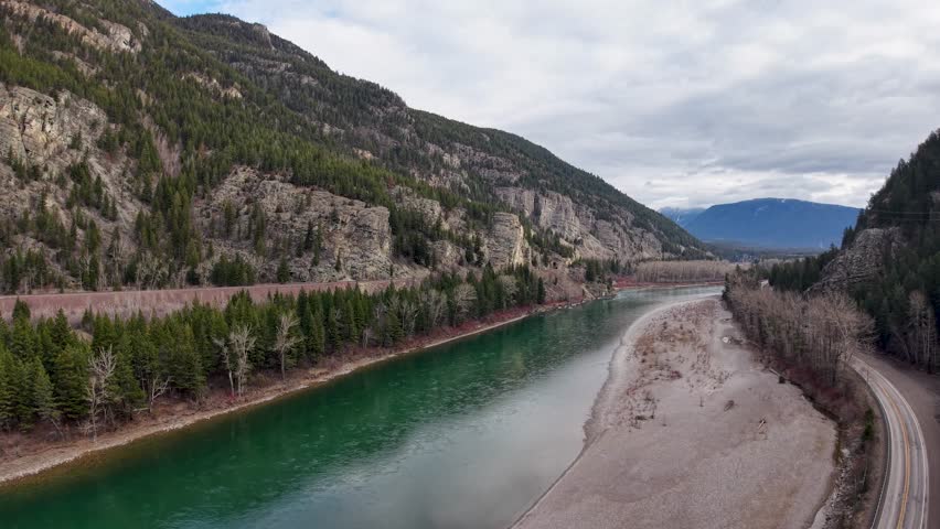 Drone aerial view of a turquoise river flowing between a rocky cliff and a wooded hillside, with a winding bank and distant mountains in the wilderness near Whitefish, Montana