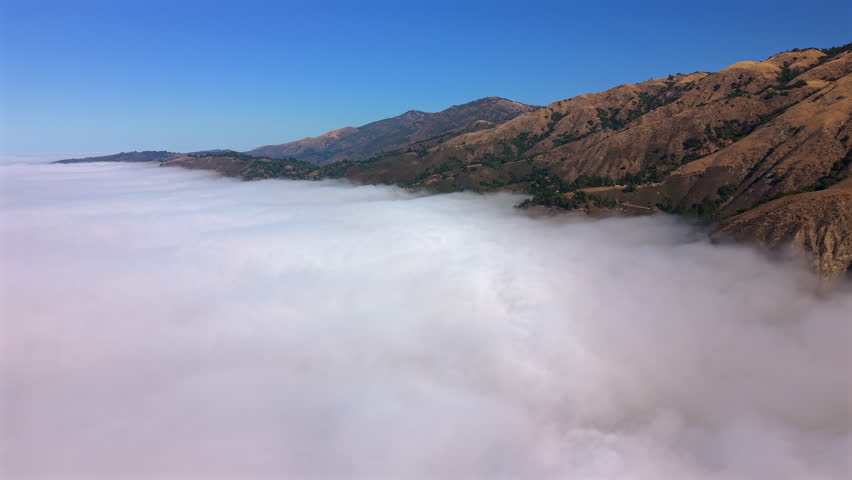 Aerial panorama of Big Sur California mountains floating above coastal fog. Pacific Ocean hidden below while rugged hills glow in warm golden light.