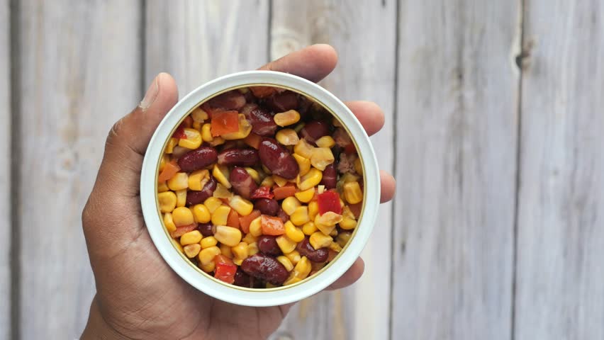 Person eating fresh mixed canned salad with corn and beans from white bowl using transparent fork