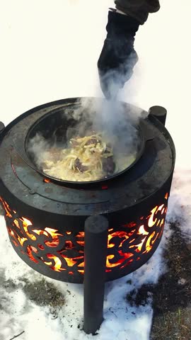 Steaming rice and meat cooking on iron stove surrounded by snow. Wilderness lifestyle, survival skills and rustic fire scene. Man stirring pilaf in hot pan outdoor, vertical shot