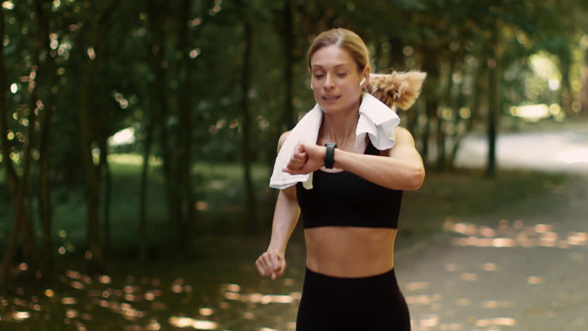Intensive cardio training. Active middle aged lady jogger running in public park and looking at her smartwatch