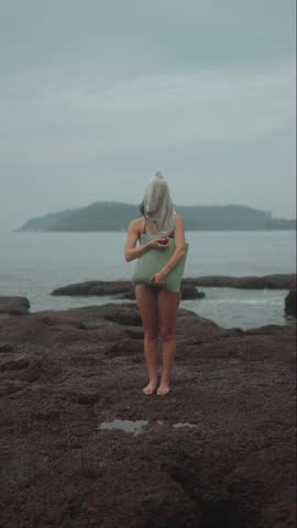 Woman stands still on rocky coast holding pillow as waves move gently behind her.