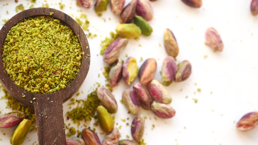Close up slow motion top view of ground pistachio nuts in wooden spoon on white background with whole pistachios scattered around