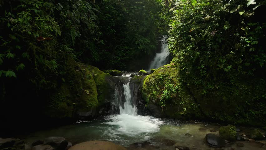 Drone flies low past multiple small cascading waterfalls dropping between moss-covered boulders into clear pools inside Mindo cloud forest, Ecuador
