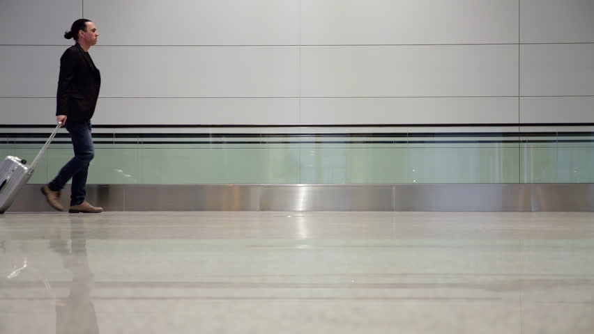 Confident man in blazer and jeans walks through a spacious, modern, and nearly empty airport terminal, pulling a small silver wheeled suitcase. Slow motion side view captures his composed stride, ready for a business trip or vacation. Empty travolator and flat wall on background