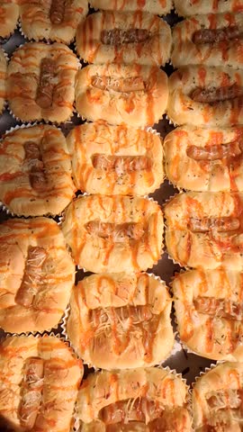Freshly baked sausage pizza breads topped with melted cheese and chili sauce are displayed in a tray at a local bakery during the day.