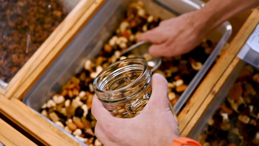 Hands scoop trail mix into glass jar at bulk food store, close up slow motion footage