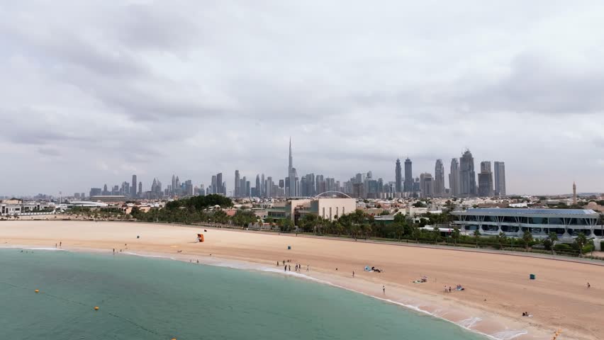 Aerial drone shot flying forward over the turquoise water and sandy shore of Kite Beach in Dubai, featuring the city skyline and Burj Khalifa.