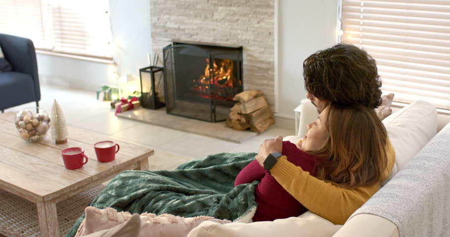 Fireplace crackling as couple is cuddling on sofa, pulling blanket and sipping red mugs for warmth. Coziness, intimacy, relaxation, warm, home, rustic, comfort