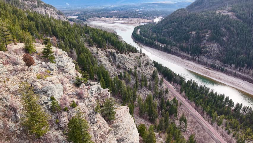 Drone view of a curving river flowing through a valley between forested hillsides and rocky slopes near Whitefish, Montana, with mountains in the distance