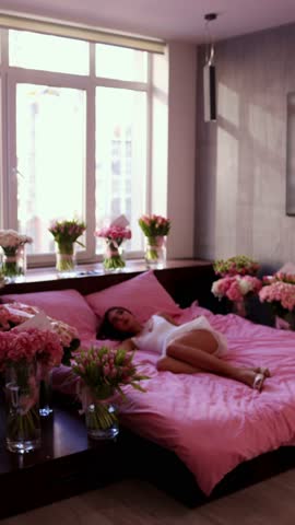 A vertical wide shot of a young woman in a white dress sitting on a pink bed, expressing joyful shock and surprise as she wakes up surrounded by dozens of flower bouquets