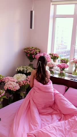 A vertical wide shot of a young woman wrapped in a pink duvet, sitting on a bed in a bright room filled with numerous pink and white flower bouquets