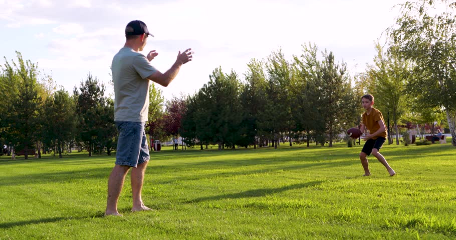 Father plays American football with son. Dad teaches American football skills in park. Family enjoys American football game on grass. Active lifestyle bonds parent and child. Summer day brings joy.