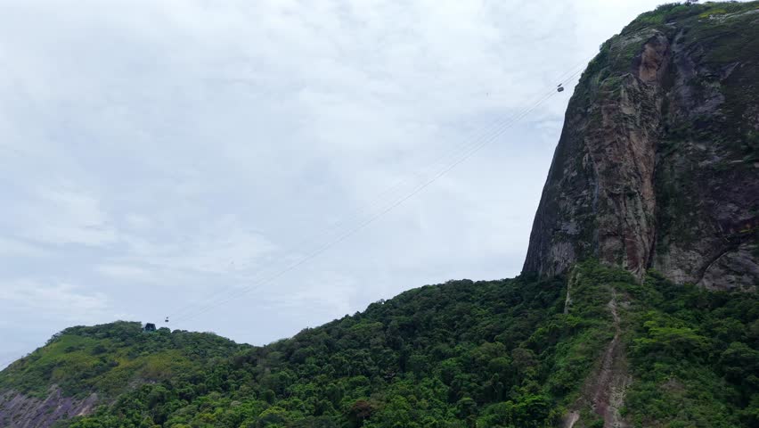 Sugarloaf Mountain towering above lush hills in Rio de Janeiro Brazil, tram transports people to top, aerial establishing pullback