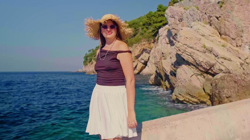 Woman on seaside pier in straw hat and white skirt looking at scenic cliffs turquoise water and green trees resort vacation mood