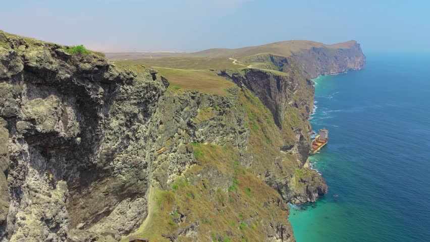 Aerial view of steep rocky cliffs and a shipwrecked cargo vessel along the turquoise coastline of the Arabian Sea near Salalah, Oman.