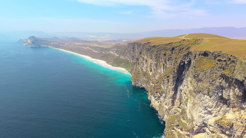 Aerial perspective of steep rocky cliffs overlooking a turquoise sea and sandy beach coastline near Salalah, Oman.