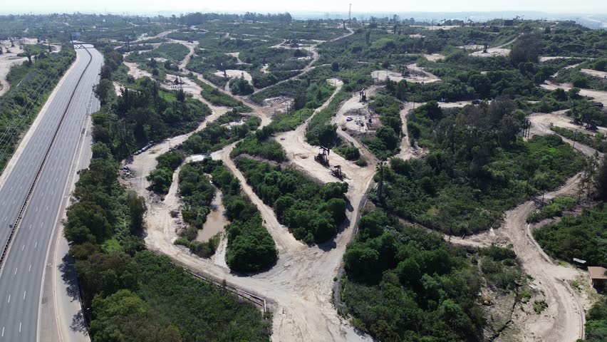 La Cienega Blvd corridor borders eastern edge of Inglewood Oil Field with visible power lines and graded slopes. Active pumpjack clusters, rolling contours, dusty service paths in Los Angeles, CA. USA