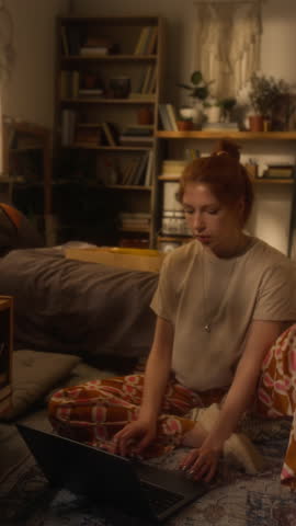 Vertical full length shot of young woman dressed in homewear using laptop while sitting on floor at home and casually working freelance, scene lit by warm lighting