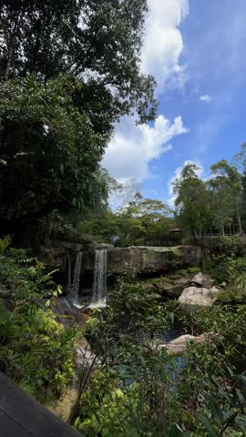 Small waterfall flowing over rocky cliff into a natural pool surrounded by lush green forest, tranquil tropical nature landscape.
