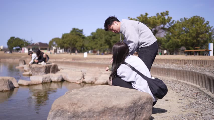 A man and a woman are sitting on a rock by a pond. The man is holding the woman