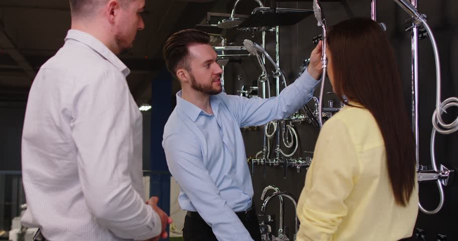 Young couple choosing shower for their bathroom standing with male friendly salesman near the showcase of plumbing shop with lots of sanitary goods. Man and woman at building market. 4k video.