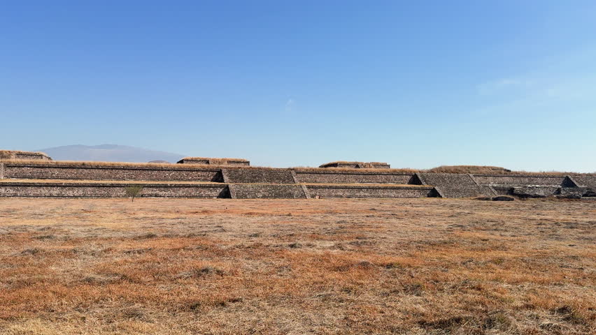 Wide shot of Teotihuacan stone terraces and plaza in Mexico under clear blue sky, with dry grass foreground and copy space for travel, history, and culture.